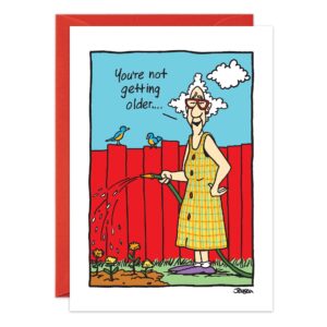 A joyful senior woman in sunglasses and a plaid dress waters her garden, framed by a red fence, blue sky, and two birds—a lively scene from "1377.jpg," the perfect playful birthday card featuring the sign: You're not getting older...