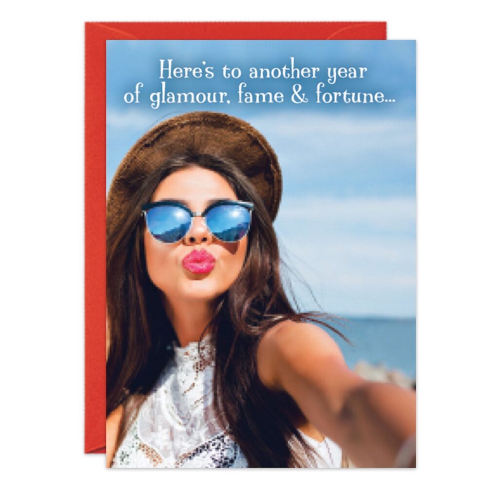 A woman in sunglasses and a hat takes a beach selfie, puckering her lips. Text reads: Here’s to another year of glamour, fame & fortune—the perfect moment for 8882.jpg.