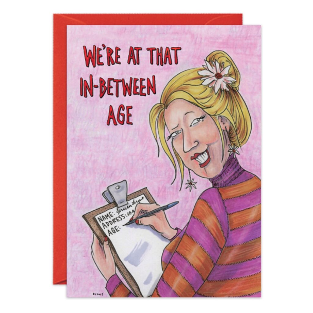 A cartoon woman with a flower in her hair grins while writing on a clipboard. "We're at that in-between age" text appears above on this playful pink birthday card (8996.jpg) featuring name, address, and age spots, with a red envelope behind.